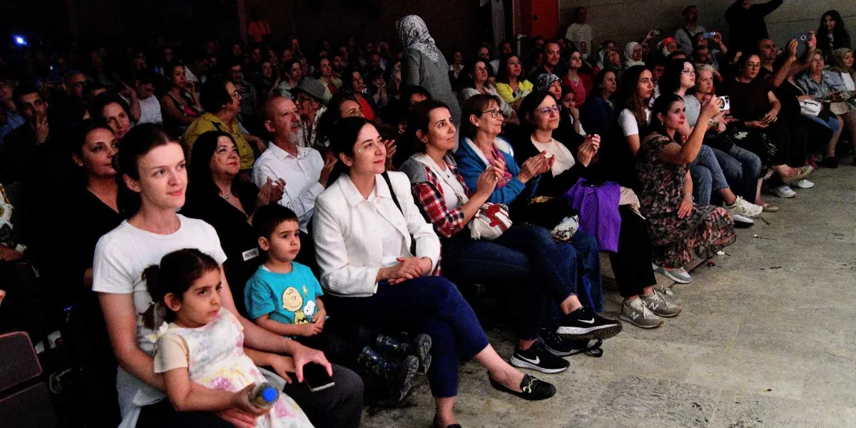 Kapalı bir mekanda, sandalyelerde yan yana oturmuş, sahnedeki gösteriyi ilgiyle izleyen ve alkışlayan kalabalık bir izleyici grubu. Ön planda kucağında küçük bir kız çocuğu tutan genç bir kadın, yanında mavi tişörtlü bir erkek çocuk ve beyaz ceketli bir kadın yer alıyor. İzleyicilerin çoğu kadınlardan oluşuyor ve birçoğu elleriyle alkış tutarak etkinliğe katılım gösteriyor.