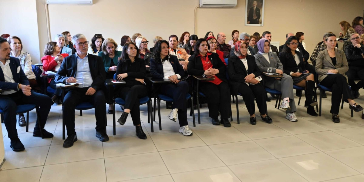 Kapalı bir salonda, sıralarda oturarak bir toplantıyı veya sunumu takip eden, ağırlıklı olarak kadınlardan oluşan kalabalık bir grup. Katılımcılar resmi ve yarı resmi kıyafetler içinde, dikkatle karşıya bakıyorlar. Salonun duvarında Mustafa Kemal Atatürk'ün bir portresi asılı.