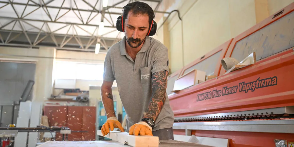 Bornova Belediyesi logolu gri bir tişört giyen, kollarında dövmeleri bulunan bir atölye çalışanı, kulaklık takmış halde endüstriyel bir kenar yapıştırma makinesinin önünde ahşap bir parçayı işliyor. Çalışan, ellerinde turuncu iş eldivenleriyle ahşabı yönlendirirken havada talaş parçacıkları uçuşuyor.