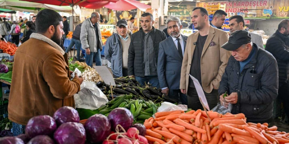 Bej renkli ceket giyen bir yetkili, beraberindeki heyetle birlikte kapalı bir pazaryerinde esnafla sohbet ediyor. Ön planda mor lahana, havuç ve salatalık gibi taze sebzelerin dizili olduğu bir tezgah yer alırken, arka planda alışveriş yapan diğer insanlar ve pazar tezgahları görülüyor.
