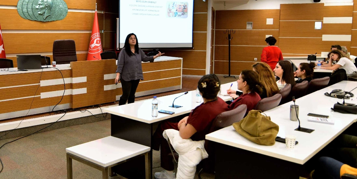 Bornova Belediyesi meclis salonunda, bir eğitmen ayakta durarak katılımcılara sunum yapıyor. Arkadaki projeksiyon perdesinde 