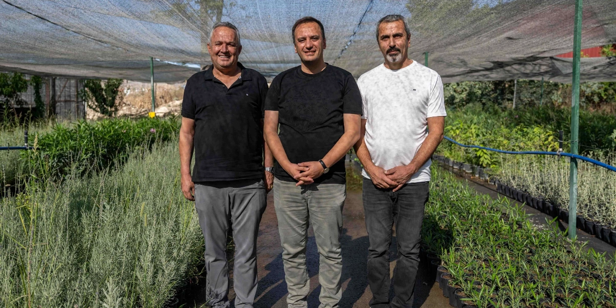 Üzeri güneşlik ile örtülü bir fidanlıkta yan yana duran üç erkek. Soldaki kişi siyah polo yaka tişört ve gri pantolon, ortadaki kişi siyah tişört ve bej pantolon, sağdaki kişi ise beyaz tişört ve koyu renk pantolon giyiyor. Arka planda saksılar içinde düzenli bir şekilde dizilmiş yeşil bitkiler ve sulama boruları yer alıyor.
