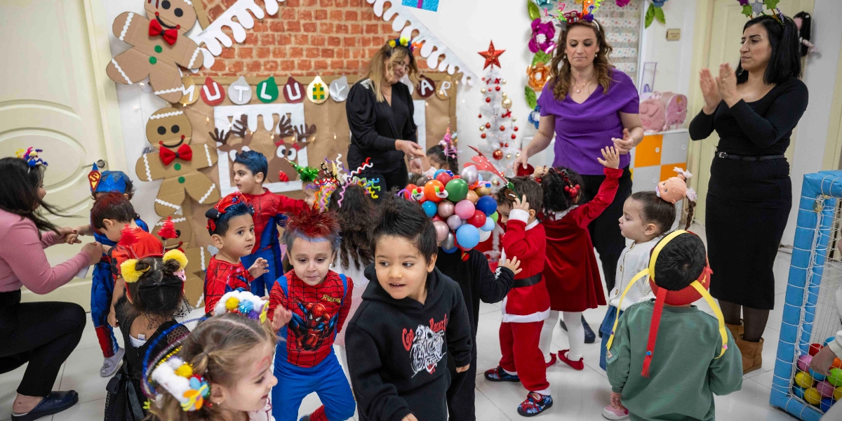 Yeni yıl kutlaması için süslenmiş bir sınıfta, kostümlü çocuklar ve öğretmenler eğleniyor. Arka planda kartondan yapılmış, üzerinde 