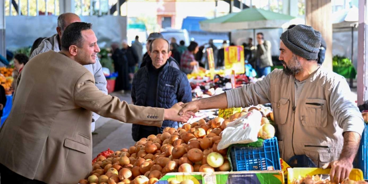 Bej renkli ceket giyen bir yetkili, kapalı bir pazaryerinde tezgahının başında duran, gri bere ve bej yelek giymiş sakallı bir esnafla el sıkışıyor. Önlerinde bol miktarda kuru soğan bulunan tezgahın arkasında, aralarında orta yaşlı bir adamın da bulunduğu birkaç kişi daha yer alıyor.