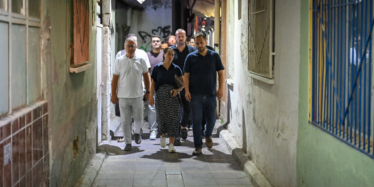 Gece vakti dar bir sokakta yürüyen, aralarında bir kadının da bulunduğu bir grup erkek, altyapı çalışmalarını incelemek üzere ilerliyor. Grup, eski binaların arasında yer alan taş döşeli yolda yan yana ve arka arkaya sıralanmış şekilde yürüyor.