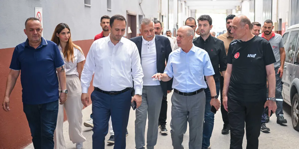 Bornova Belediye Başkanı Ömer Eşki ve beraberindeki heyet, bir yangın bölgesinde incelemelerde bulunurken yürürken görülüyor. Heyette, üzerinde AFAD yazılı siyah tişört giyen bir yetkilinin yanı sıra çeşitli belediye çalışanları ve görevliler yer alıyor. Grup, gri duvarlı bir koridorda ilerleyerek durum değerlendirmesi yapıyor.