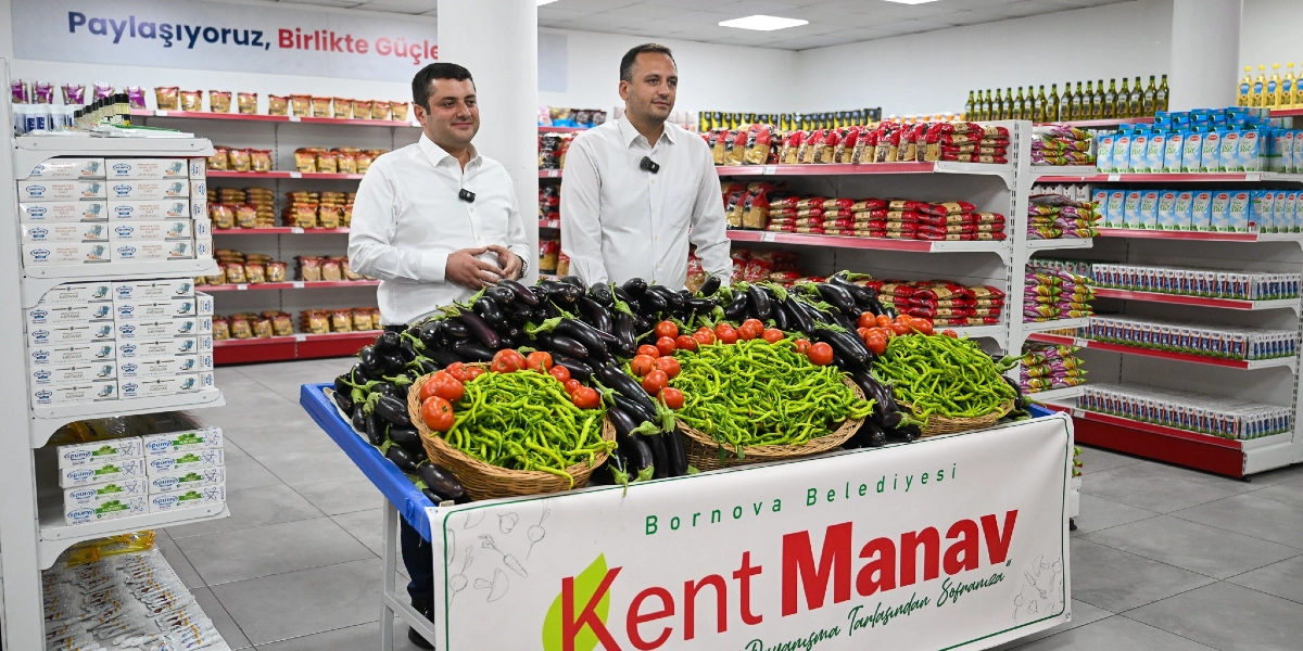 Beyaz gömlekli iki erkek, Bornova Belediyesi'ne ait Kent Manav reyonunun önünde duruyor. Önlerindeki stantta sepetler içinde taze patlıcan, domates ve yeşil biberler sergileniyor. Arka planda ise raflara düzenli bir şekilde yerleştirilmiş gıda ürünleri ve duvar üzerinde 