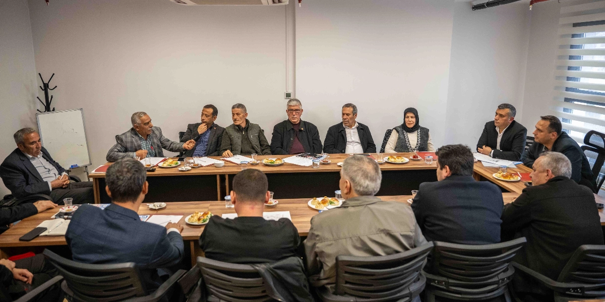 U-şeklindeki bir toplantı masasının etrafında toplanmış, aralarında Başkan Ömer Eşki'nin de bulunduğu bir grup muhtar ve yerel yetkili. Katılımcılar resmi kıyafetlerle oturmuş, önlerinde evraklar ve ikramlık tabaklar bulunan masada görüşme yapıyorlar. Arka planda beyaz bir duvar ve sağ tarafta jaluzili bir pencere bulunuyor.