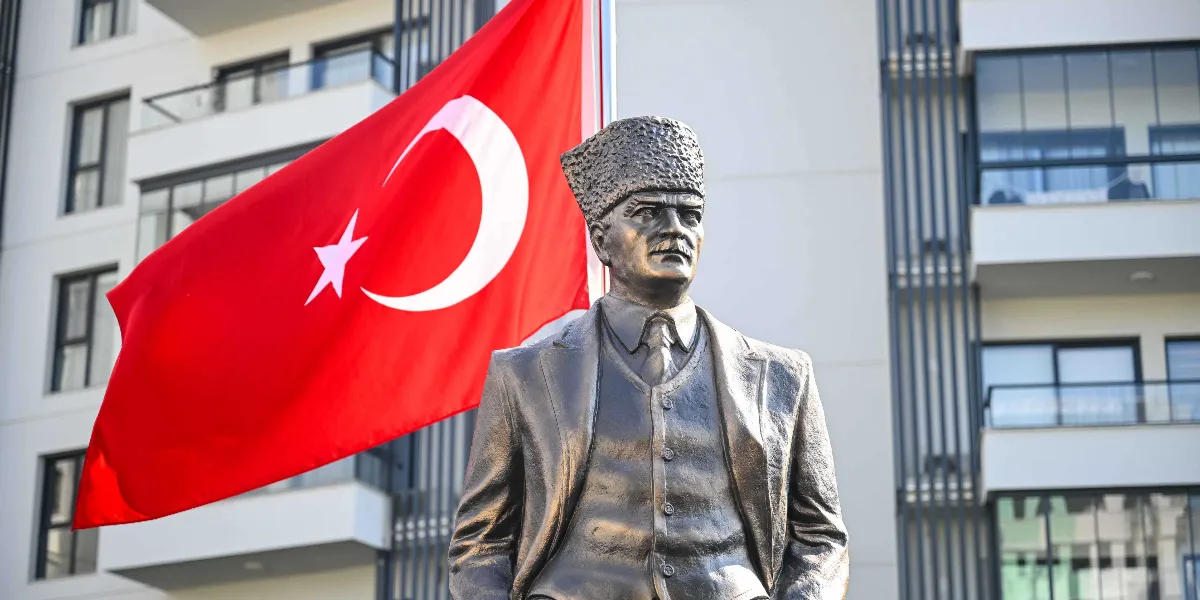 Modern bir apartman binasının önünde yer alan, kalpaklı Mustafa Kemal Atatürk büstü ve arkasında dalgalanan büyük bir Türk bayrağı.