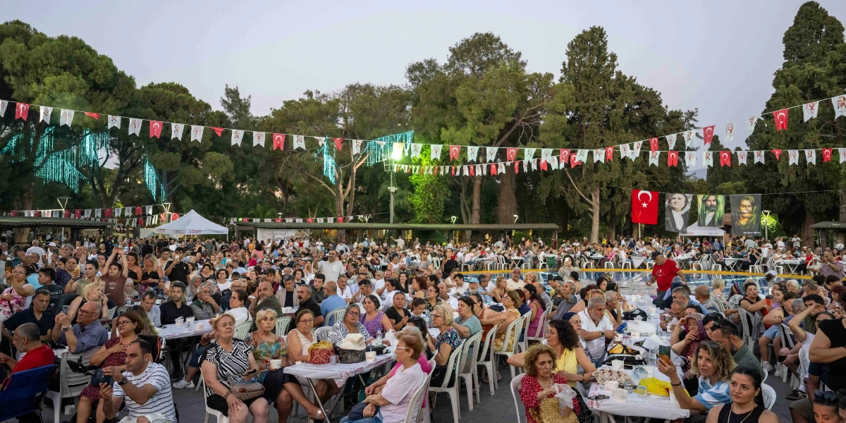 Açık havada, ağaçlarla çevrili bir park alanında kurulan uzun masalarda iftar yemeği için toplanmış kalabalık bir grup insan. Masaların üzeri beyaz örtülerle kaplı ve üzerinde yemek kapları bulunuyor. Alanın üst kısmı, Türk bayrakları ve üzerinde siyasi figürlerin görsellerinin bulunduğu flamalarla süslenmiş. Arka planda, ağaçların arasına asılmış büyük boyutta Atatürk, Hacı Bektaş Veli ve Pir Sultan Abdal portreleri ile Türk bayrağı yer alıyor. İnsanlar masalarda oturmuş sohbet ederken, bazıları telefonlarıyla fotoğraf çekiyor.