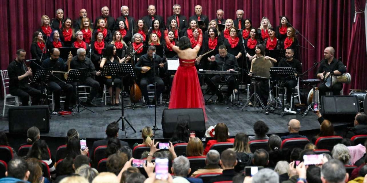 Kırmızı perdeli bir sahnede, siyah kıyafetler giymiş kalabalık bir koro ve orkestra grubu performans sergiliyor. Grubun önünde, sırtı izleyicilere dönük, kırmızı uzun bir elbise giymiş kadın şef orkestrayı yönetiyor. Müzisyenler arasında bağlama, klavye ve vurmalı çalgılar çalanlar bulunuyor. Salonu dolduran izleyiciler, sahnedeki performansı cep telefonlarıyla kaydederek izliyor.