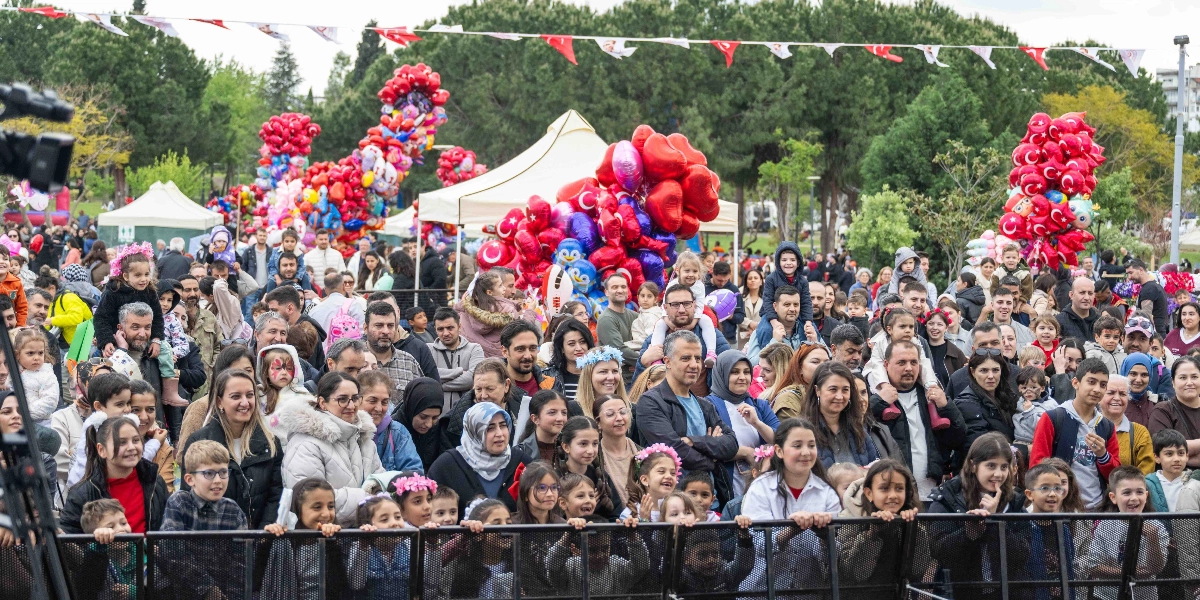 Açık havada düzenlenen bir etkinlikte, metal bariyerlerin arkasında duran ve sahneye bakan kalabalık bir çocuk ve yetişkin grubu. Arka planda ağaçlar, beyaz çadırlar ve üzerinde Türk bayrakları bulunan kırmızı kalp şeklinde balon demetleri yer alıyor. Etkinliğe katılan çocuklar ve aileleri neşeli bir atmosferde bir arada bulunuyor.