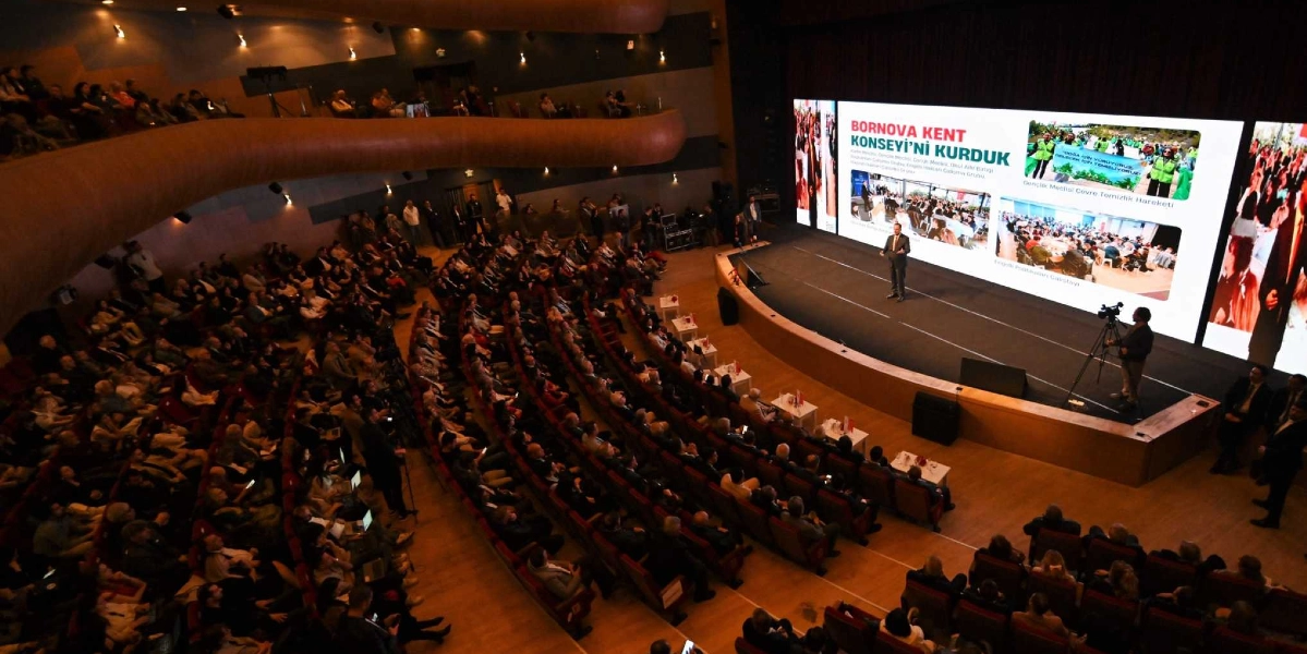 Modern bir konferans salonunda, sahnedeki dev ekranda Bornova Kent Konseyi'nin iki yıllık faaliyetlerinin sunulduğu bir etkinlik. Sahnenin merkezinde bir konuşmacı yer alırken, dev ekranda 