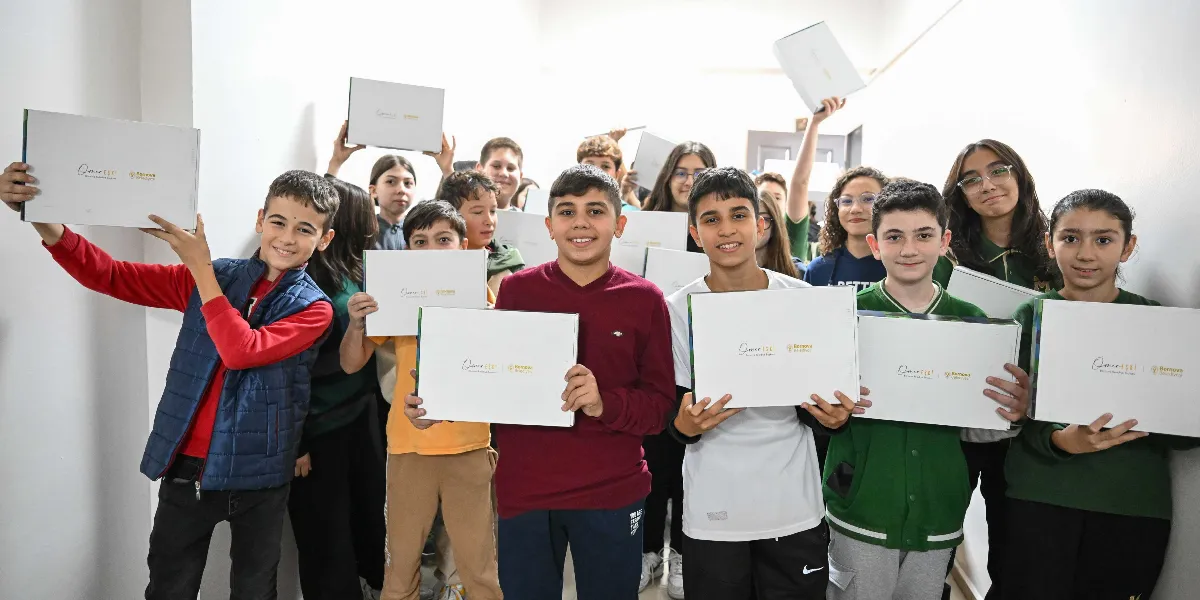 Beyaz bir koridorda bir araya gelmiş, ellerinde üzerinde BELGEM logosu bulunan beyaz hediye kutularını gülümseyerek havaya kaldıran veya önlerinde tutan bir grup öğrenci.
