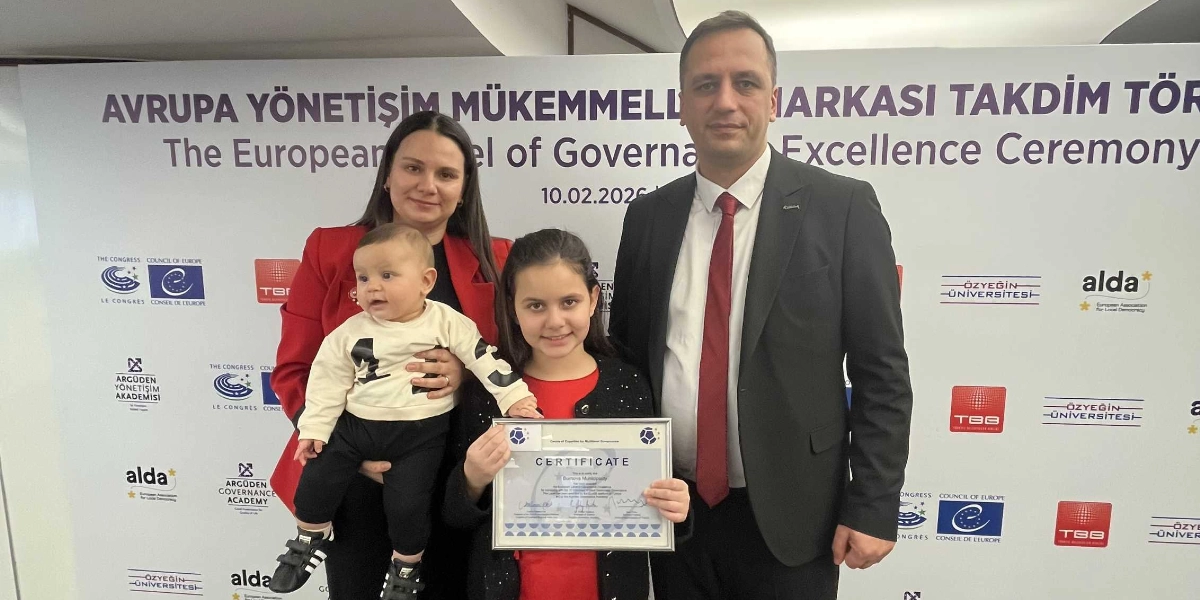 Avrupa Yönetişim Mükemmellik Markası Takdim Töreni'nde, elinde sertifika tutan bir kız çocuğu, kucağında bebek olan bir kadın ve yanında duran takım elbiseli bir erkek yan yana poz veriyor. Arka planda törenin adının ve çeşitli kurumların logolarının yer aldığı beyaz bir pano bulunuyor.
