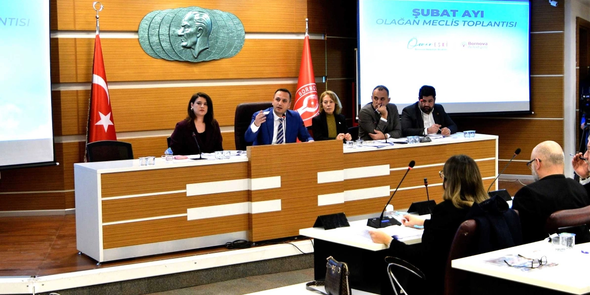 Bornova Belediyesi Şubat Ayı Olağan Meclis Toplantısı'nda, başkanlık divanında oturan yetkililer konuşma yaparken, meclis üyeleri masalarda oturarak toplantıyı takip ediyor. Arka planda duvarda Atatürk kabartması, iki yanda Türk bayrağı ve Bornova Belediyesi flaması yer alıyor. Sağ taraftaki projeksiyon ekranında 