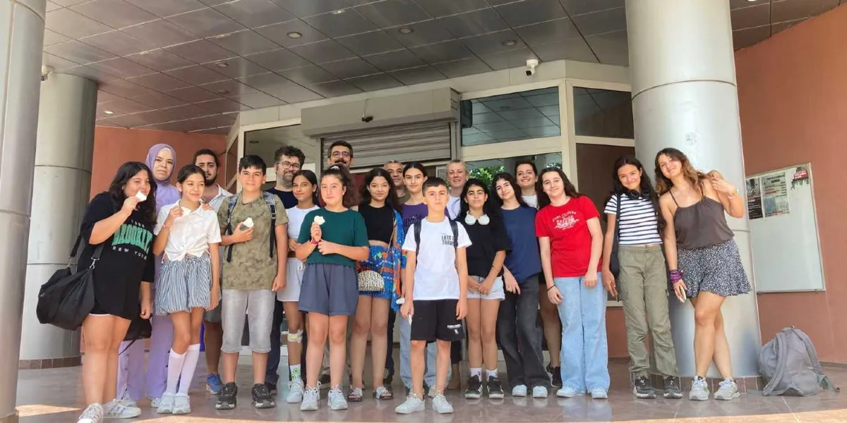 Bornova'da bir binanın girişinde yan yana duran, aralarında yetişkinlerin de bulunduğu, çoğunluğu genç ve çocuklardan oluşan kalabalık bir grup. Gruptaki kişiler günlük kıyafetler içinde, gülümseyerek kameraya bakıyor. Bazı gençler ellerinde küçük kağıtlar tutuyor ve grubun yanında bir sırt çantası duruyor.