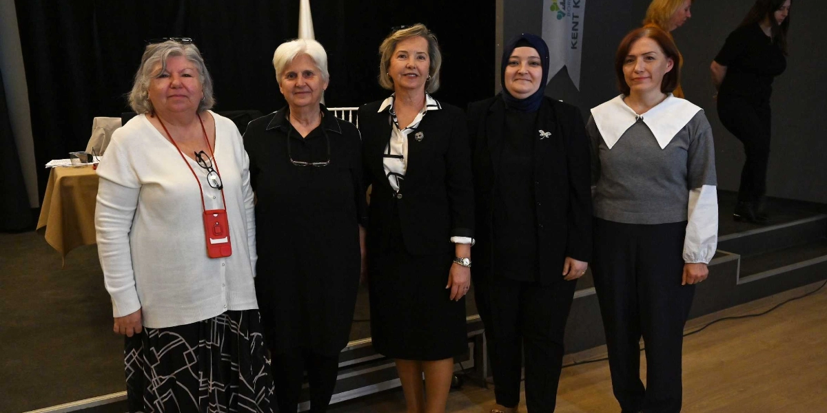 Kapalı bir mekanda, sahne önünde yan yana duran beş kadın. Soldan sağa doğru; beyaz hırkalı ve desenli etekli, siyah elbiseli, siyah ceket ve beyaz gömlekli, siyah kıyafetli ve başörtülü, gri kazaklı ve beyaz yakalı bir kadın yer alıyor. Arka planda siyah bir perde ve Bornova Kent Konseyi yazılı bir flama bulunuyor.