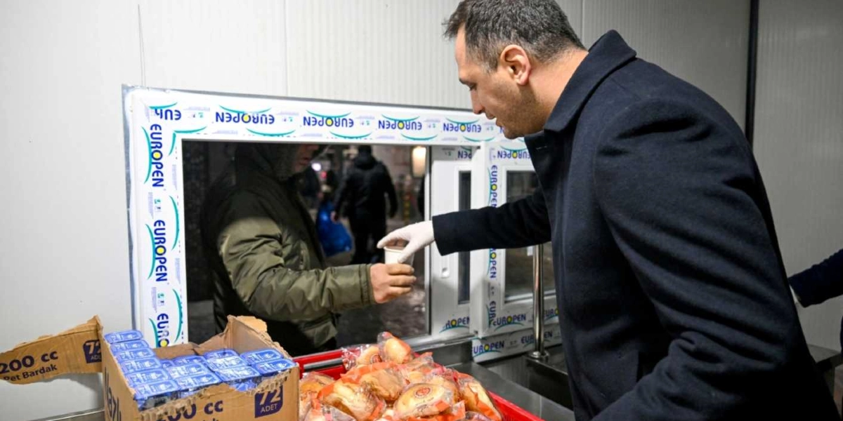 Koyu renk palto giymiş bir erkek, beyaz eldivenli eliyle bir pencereden dışarıdaki kişiye sıcak içecek ikram ediyor. Pencerenin önündeki tezgahta paketlenmiş poğaçalar ve karton bardak kutuları bulunuyor.