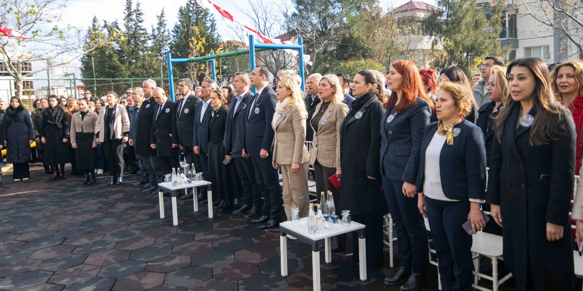 Açık havada, park zemininde yan yana dizilmiş bir grup kadın ve erkek, resmi bir tören için ayakta duruyor. Çoğunluğu koyu renkli kıyafetler giymiş olan katılımcıların yakalarında anma etkinliğine dair küçük rozetler bulunuyor. Arka planda çocuk parkı oyun grubu ve ağaçlar görülürken, tören alanındaki katılımcılar ciddiyetle karşıya bakıyor.