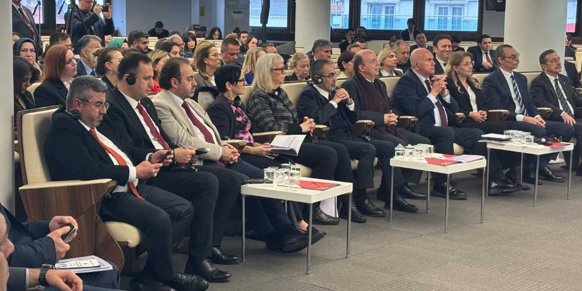 Resmi bir toplantı salonunda, takım elbiseli ve resmi kıyafetli kişilerin yan yana dizilmiş koltuklarda oturduğu bir konferans ortamı. Katılımcıların çoğu dikkatle dinlerken, bazıları önlerindeki küçük masalarda bulunan belgeleri inceliyor veya not alıyor. Salonun genelinde ciddi bir atmosfer hakimken, katılımcılar arasında hem erkekler hem de kadınlar bulunuyor. Ön planda, kulaklık takan ve toplantıyı takip eden bir grup katılımcı net bir şekilde görülüyor.