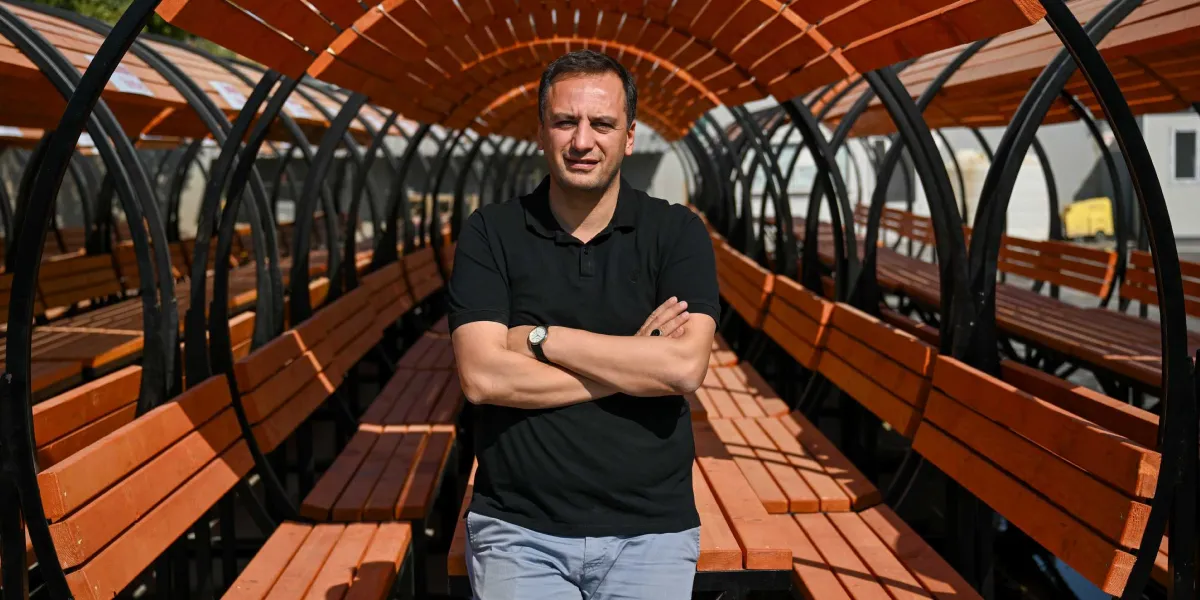Siyah polo yaka tişört giymiş bir erkek, kollarını kavuşturmuş şekilde, ahşap çıtalardan yapılmış tünel biçimindeki modern bir oturma alanının ortasında duruyor. Siyah metal iskeletli ve ahşap oturaklı bu yapı, parklarda kullanılmak üzere tasarlanmış yeni nesil bir dinlenme alanını temsil ediyor.