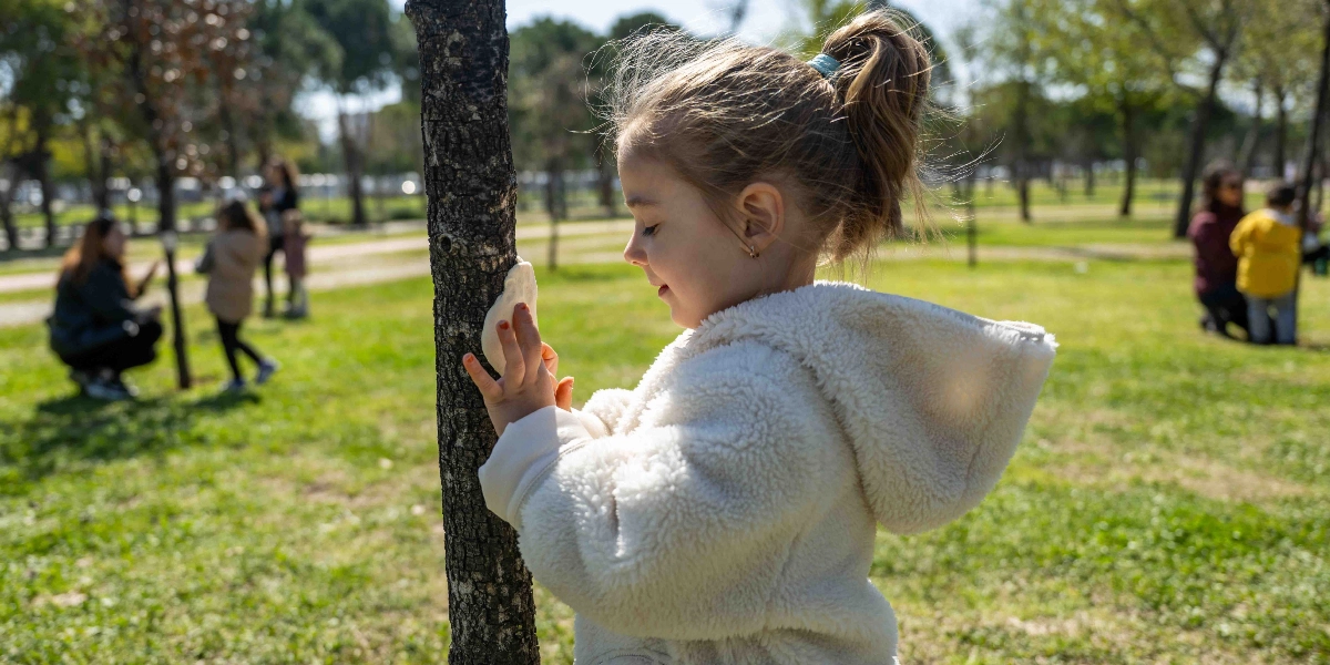 Beyaz, peluş bir mont giymiş küçük bir kız çocuğu, güneşli bir parkta ağaç gövdesine oyun hamuru benzeri beyaz bir maddeyi yapıştırarak dikkatle inceliyor. Arka planda çimenlik alanda vakit geçiren diğer çocuklar ve yetişkinler yer alıyor.