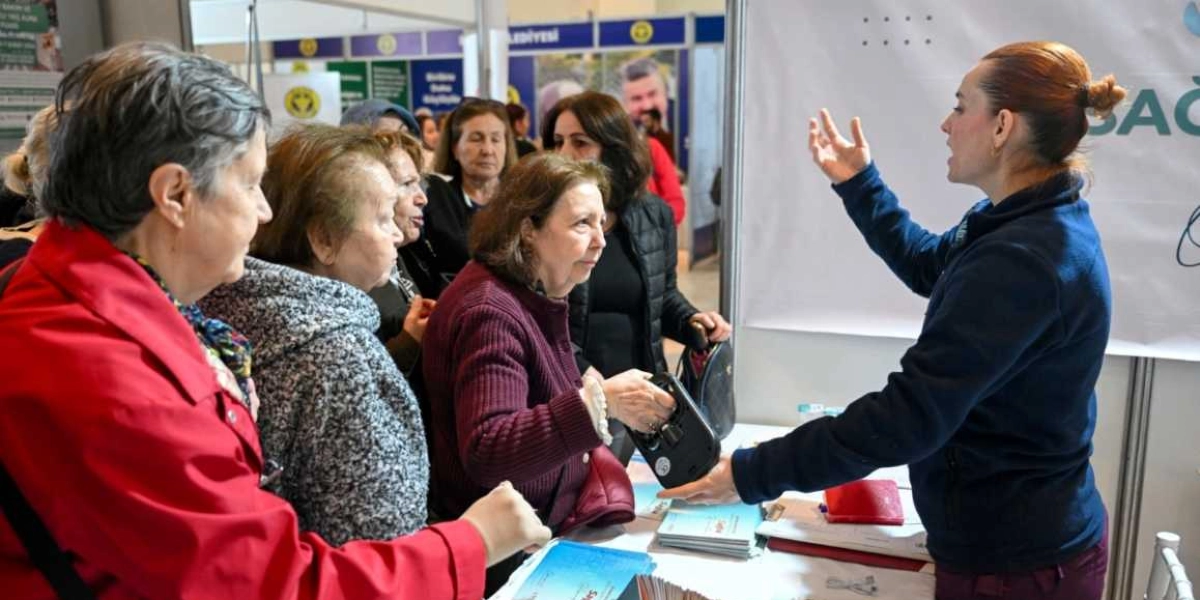 Kapalı bir alanda kurulan stantta, görevli bir kadın elindeki cihazla yaşlı bir kadına sağlık taraması veya bilgilendirme yapıyor. Görevli kadının karşısında, sırasını bekleyen veya süreci izleyen birkaç kadın daha bulunuyor. Masanın üzerinde broşürler ve çeşitli sağlık ekipmanları yer alıyor. Arka planda Bornova Belediyesi'ne ait olduğu anlaşılan logolu panolar ve bilgilendirme afişleri görülüyor.