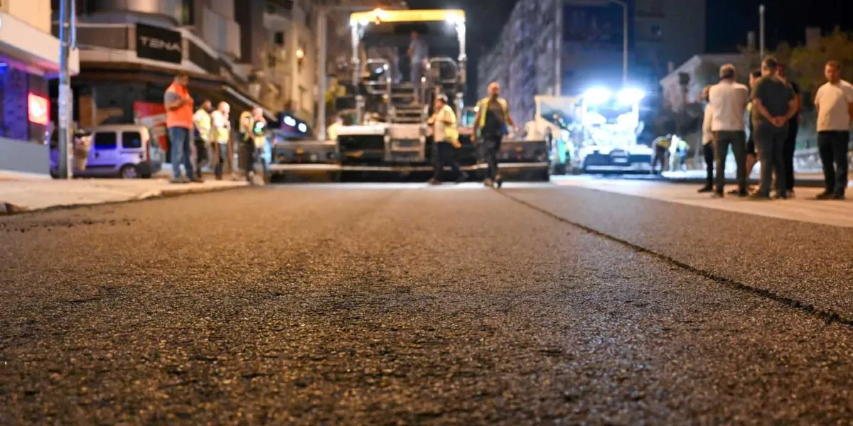 Gece saatlerinde Mustafa Kemal Caddesi'nde devam eden asfalt serme çalışması. Yolun ortasında büyük bir asfalt serme makinesi bulunurken, çevresinde reflektörlü yelek giyen işçiler ve çalışmaları izleyen insanlar yer alıyor. Çalışma alanı, iş makinelerinin güçlü ışıklarıyla aydınlatılıyor.