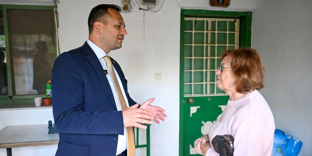 Lacivert takım elbiseli ve bej kravatlı bir erkek, beyaz duvarlı ve yeşil kapılı bir evin önünde, gözlüklü ve pembe kazaklı yaşlı bir kadınla sohbet ediyor.