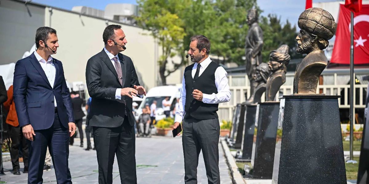Açık havada, yan yana dizilmiş tarihi figürlerin büstlerinin bulunduğu bir alanda, yakalarına mikrofon takılı üç erkek konuşuyor. Soldaki iki kişi koyu renk takım elbiseli, sağdaki kişi ise beyaz gömlek ve siyah yelek giymiş durumda. Arka planda Türk bayrağı ve ağaçlar görülüyor.