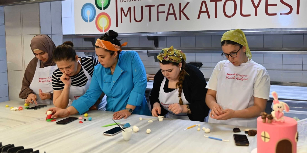 Bornova Belediyesi Mutfak Atölyesi'nde önlük giymiş beş kadın, uzun bir mutfak tezgahı etrafında toplanmış şeker hamuruyla figürler hazırlıyor. Mavi şef ceketi giyen bir eğitmen, diğer katılımcılara şeker hamuru şekillendirme konusunda rehberlik ediyor. Tezgahın üzerinde küçük hamur parçaları, modelleme araçları ve telefonlar bulunurken, sağ tarafta üzerinde tavşan figürü olan pembe bir pasta yer alıyor. Arka planda ise üzerinde 