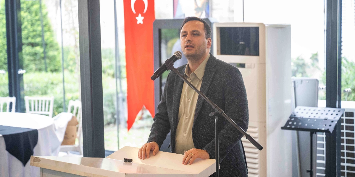 Koyu renkli bir ceket ve açık renkli bir gömlek giymiş bir erkek, kürsüde mikrofon başında konuşma yapıyor. Arka planda dikey bir Türk bayrağı, beyaz bir klima ve cam kenarında beyaz sandalyeli masalar yer alıyor.