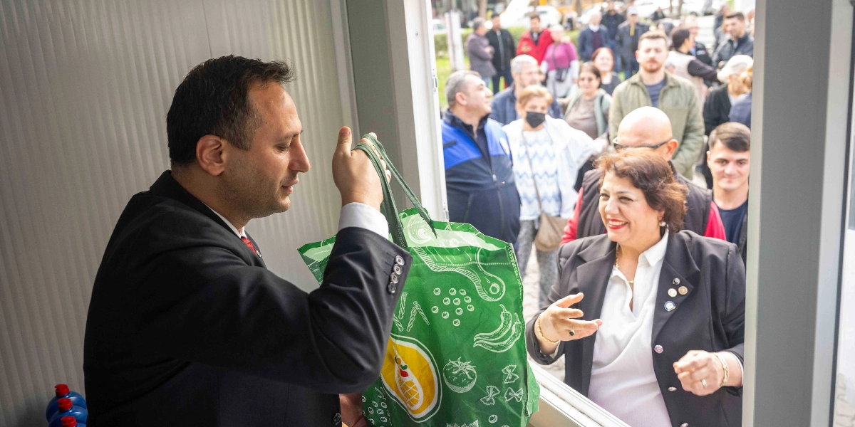 Koyu renk takım elbiseli bir erkek, üzerinde sebze ve makarna desenleri bulunan yeşil bez bir çantayı, dışarıda bekleyen güler yüzlü bir kadına uzatıyor. Kadın, beyaz gömlek ve siyah ceket giymiş, elini uzatarak çantayı alıyor. İkilinin arkasında, Atık Bank Projesi'nin açılışına katılan kalabalık bir grup insan bekliyor.