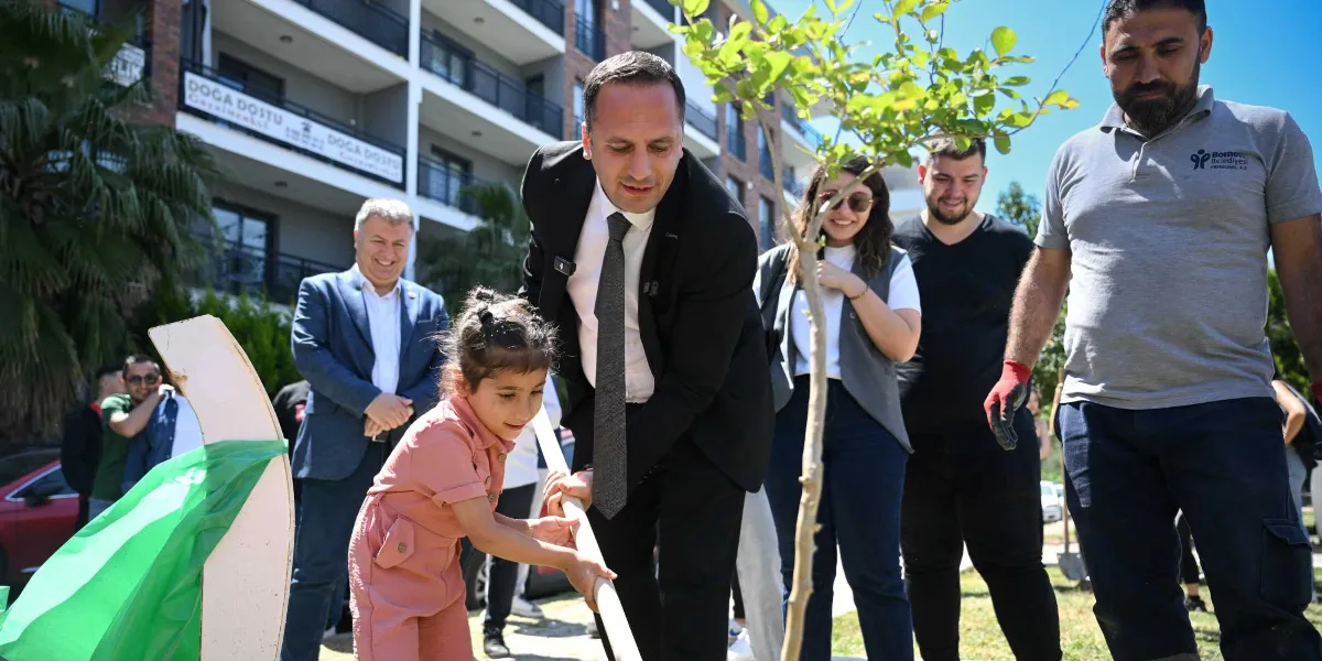 Bornova Belediye Başkanı ve beraberindekiler, bir çocukla birlikte fidan dikimi etkinliği gerçekleştiriyor. Başkan, takım elbisesiyle fidanın destek çubuğunu tutarken, pembe tulumlu bir kız çocuğu da ona eşlik ediyor. Arka planda belediye personeli ve vatandaşlar gülümseyerek onları izliyor. Etkinlik, üzerinde 