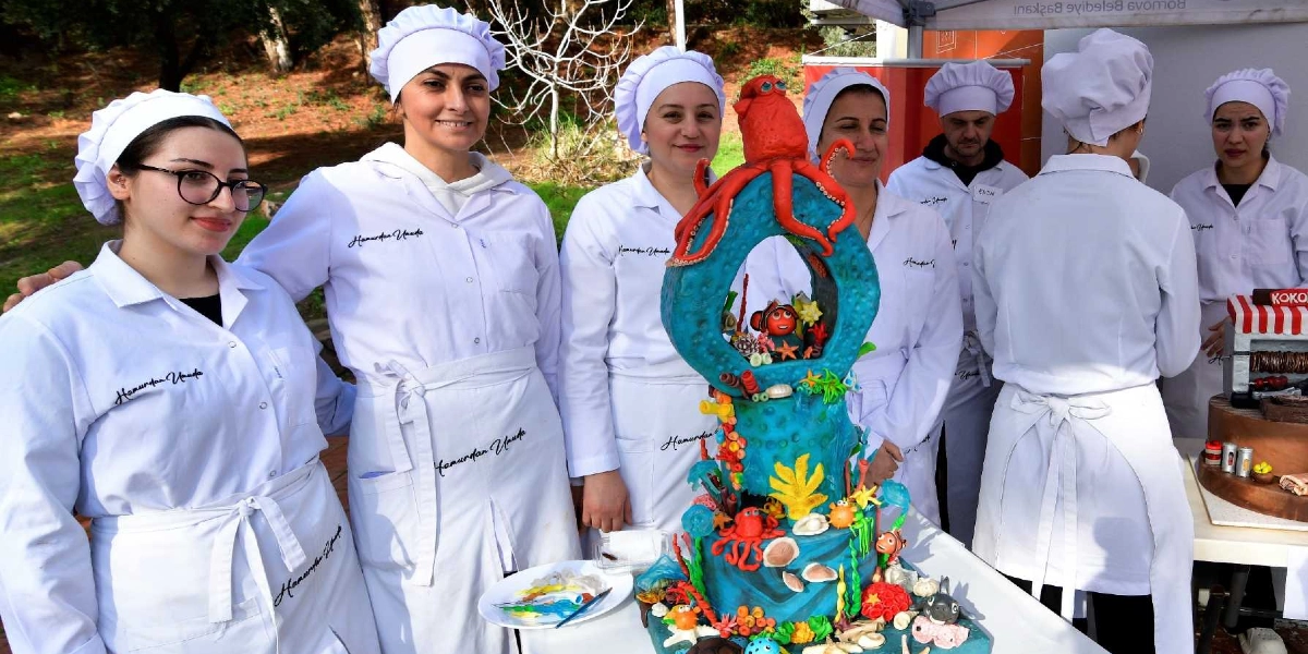 Beyaz şef önlüğü ve kepi giymiş bir grup kadın ve erkek, dış mekanda kurulan bir stantta yan yana duruyor. Önlerinde, üzerinde deniz temalı figürlerin ve tepesinde kırmızı bir ahtapotun bulunduğu, su altı dünyasını andıran renkli ve detaylı bir pasta sergileniyor. Önlüklerin üzerinde 