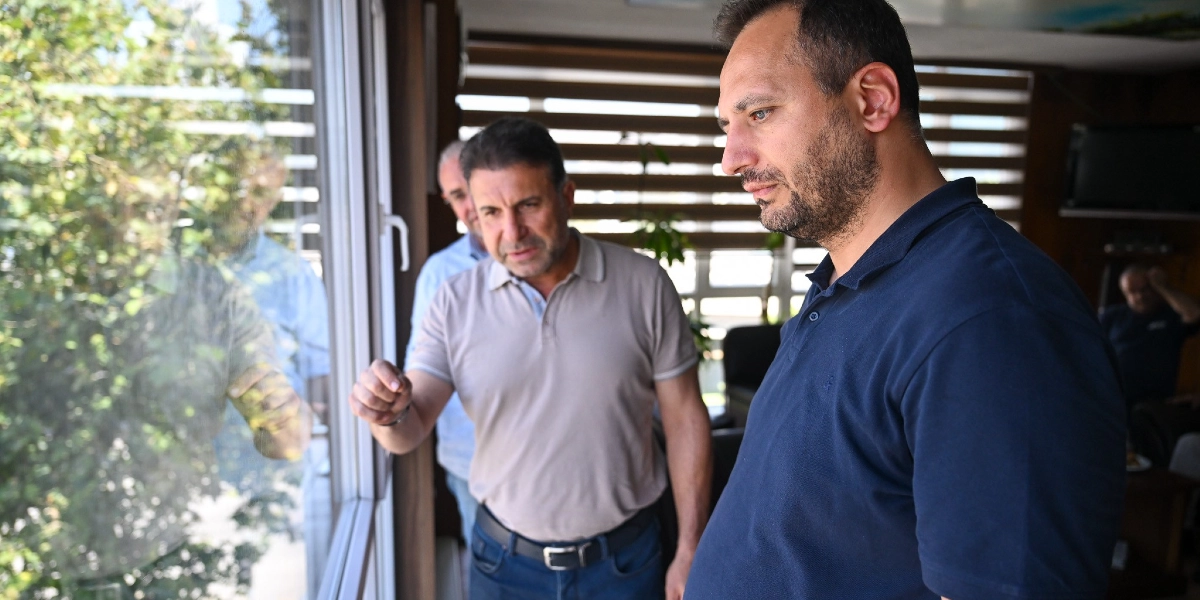 Bej polo yaka tişörtlü bir adam, pencereden dışarıyı işaret ederek yanında duran lacivert polo yaka tişörtlü adama bir şeyler anlatıyor. Arka planda ise başka bir kişi daha bulunuyor. Üçlü, iç mekanda pencere kenarında bir arada duruyor.