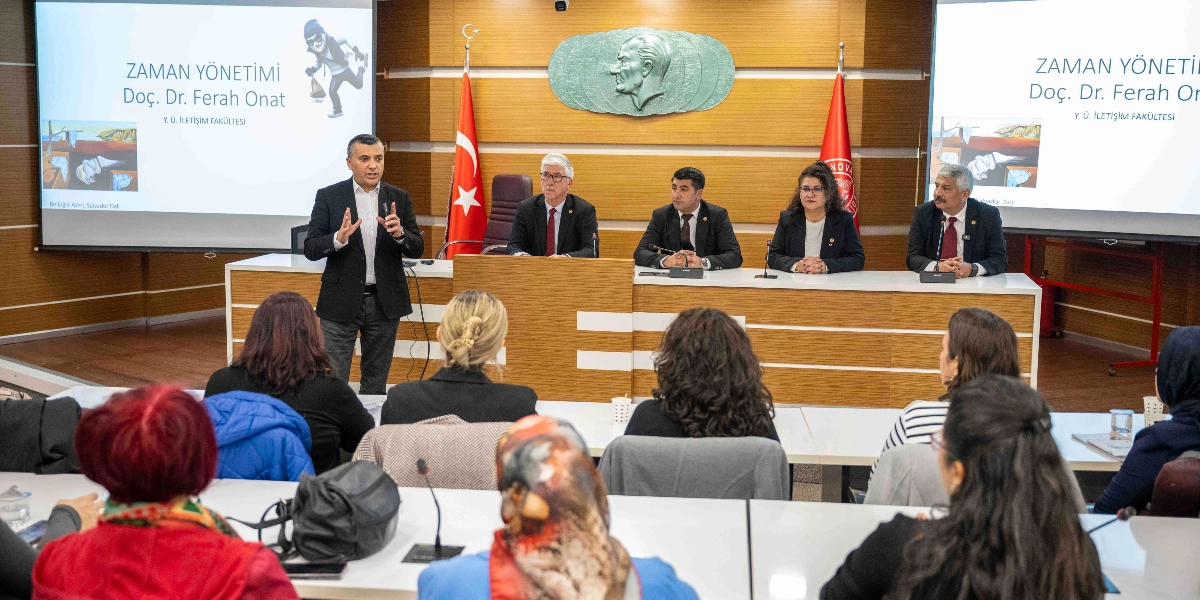 Bornova Muhtarlar Akademisi'nde düzenlenen eğitim toplantısında, bir konuşmacı ayakta sunum yaparken, masada oturan dört kişilik bir heyet ve onları dinleyen muhtarlar yer alıyor. Arka plandaki projeksiyon ekranlarında 
