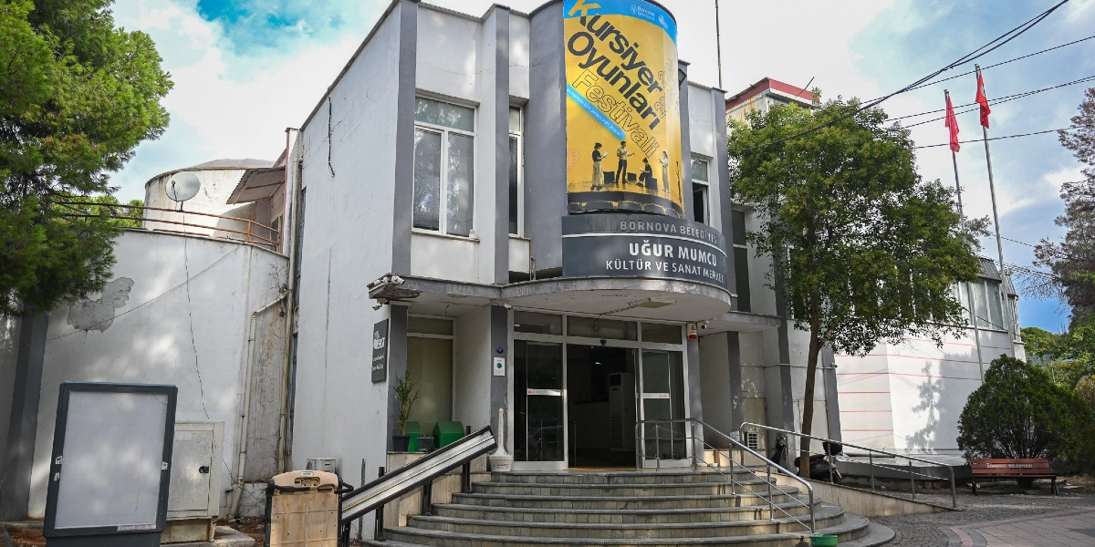 Bornova Belediyesi Uğur Mumcu Kültür ve Sanat Merkezi binasının dış cephesi. Binanın girişinde yarım daire şeklinde yükselen sarı renkli bir afişte Kursiyer Oyunları Festivali yazısı ve üzerinde tiyatro yapan insanların silüetleri yer alıyor. Binanın girişinde basamaklı bir merdiven ve engelli erişimi için bir rampa bulunuyor. Binanın sağ tarafında iki adet Türk bayrağı dalgalanıyor.