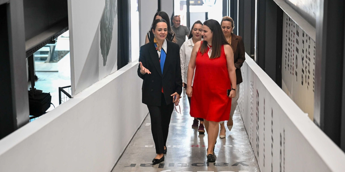 Modern bir binanın beyaz duvarlı, uzun koridorunda yürüyen bir grup kadın. Önde, siyah ceket ve pantolon giyen bir kadın el hareketleriyle konuşurken, yanında kırmızı kolsuz bir elbise giyen başka bir kadın ona eşlik ediyor. Arkalarında ise onları takip eden diğer kadınlar ve bir erkek yer alıyor. Koridorun zemininde büyük harflerle yazılmış metinler bulunuyor.