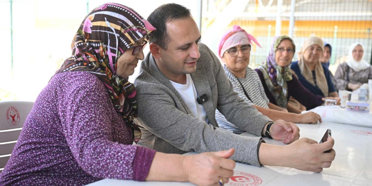 Gri ceketli bir erkek, Bornova Belediyesi logolu beyaz plastik masada oturan başörtülü yaşlı bir kadınla birlikte akıllı telefondan bir şeye bakıyor. Masanın devamında yan yana oturan diğer kadınlar da onlara eşlik ediyor ve hep birlikte gülümseyerek sohbet ediyorlar.