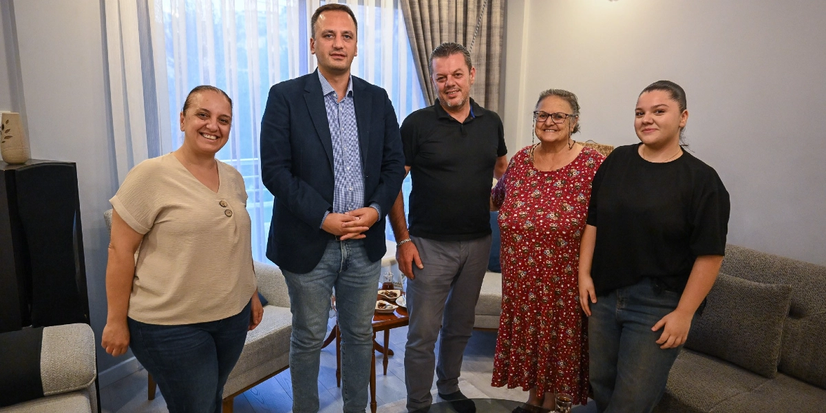 Beş kişilik bir grup, ev ortamında yan yana durarak objektife gülümsüyor. Grubun merkezinde, lacivert ceket ve mavi gömlek giymiş bir erkek yer alıyor. Onun solunda bej renkli bir bluz ve kot pantolon giyen bir kadın, sağında ise siyah polo yaka tişörtlü bir erkek, çiçek desenli uzun bir elbise giyen orta yaşlı bir kadın ve siyah tişörtlü genç bir kadın bulunuyor. Arka planda perdeler ve oturma grubu yer alıyor.