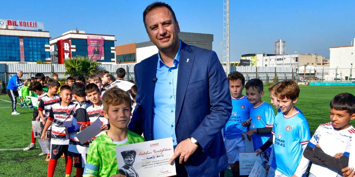 Açık havada, yeşil bir futbol sahasında, lacivert ceketli ve açık mavi gömlekli bir yetişkin, elinde tuttuğu katılım sertifikasını yeşil formalı bir çocuğa veriyor. Çocuğun elindeki sertifikanın üzerinde Atatürk fotoğrafı ve 