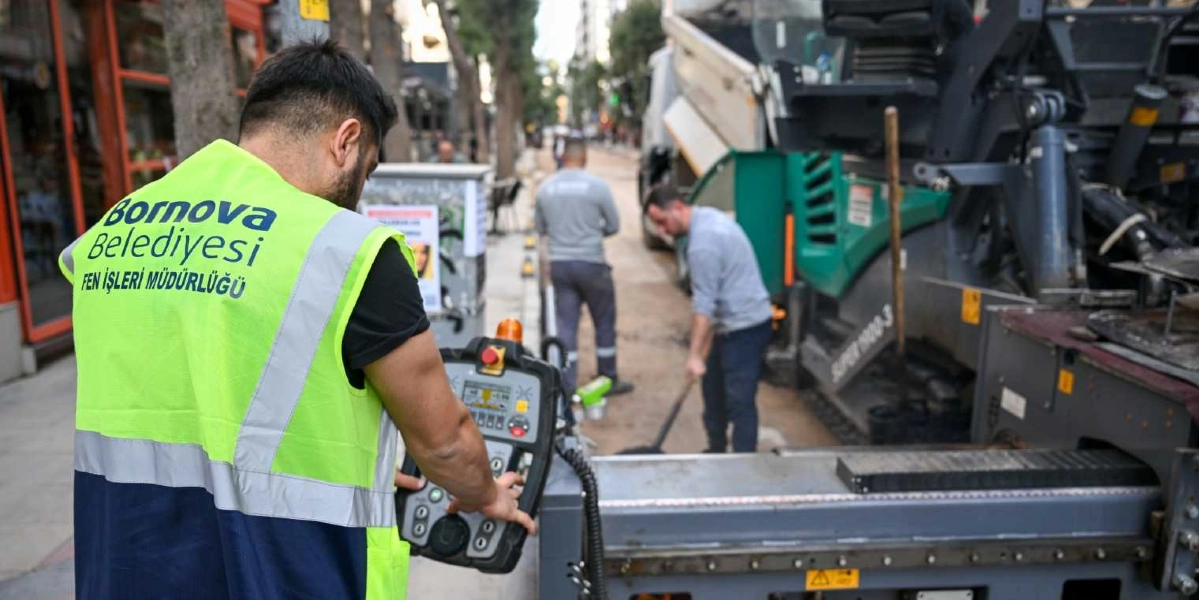 Bornova Belediyesi Fen İşleri Müdürlüğü yeleği giymiş bir görevli, elindeki uzaktan kumanda paneliyle yol asfaltlama makinesini yönetiyor. Arka planda diğer belediye çalışanları yol yenileme çalışmalarına devam ediyor.