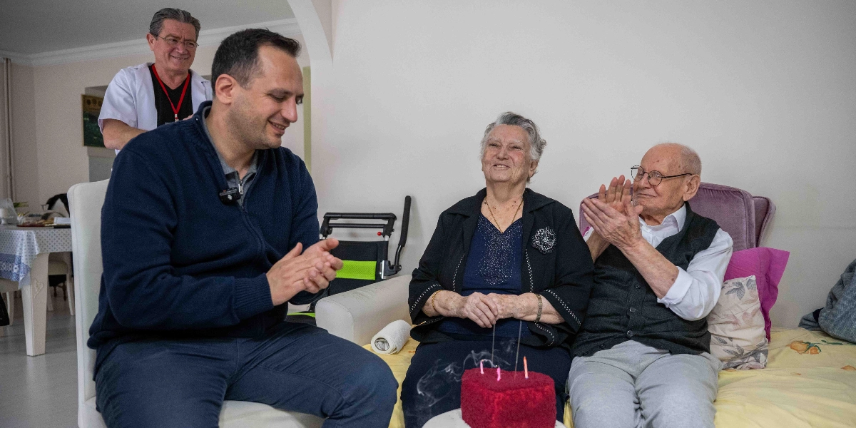 Ev ortamında, yaşlı bir çiftin önünde kalp şeklinde kırmızı bir pasta dururken, yanlarında oturan bir erkek ve arkalarında duran bir görevli alkışlayarak onlara eşlik ediyor. Yaşlı çift gülümseyerek pastaya bakarken, erkek misafir neşeyle ellerini çırpıyor, arkadaki görevli ise gülümseyerek bu anı izliyor.