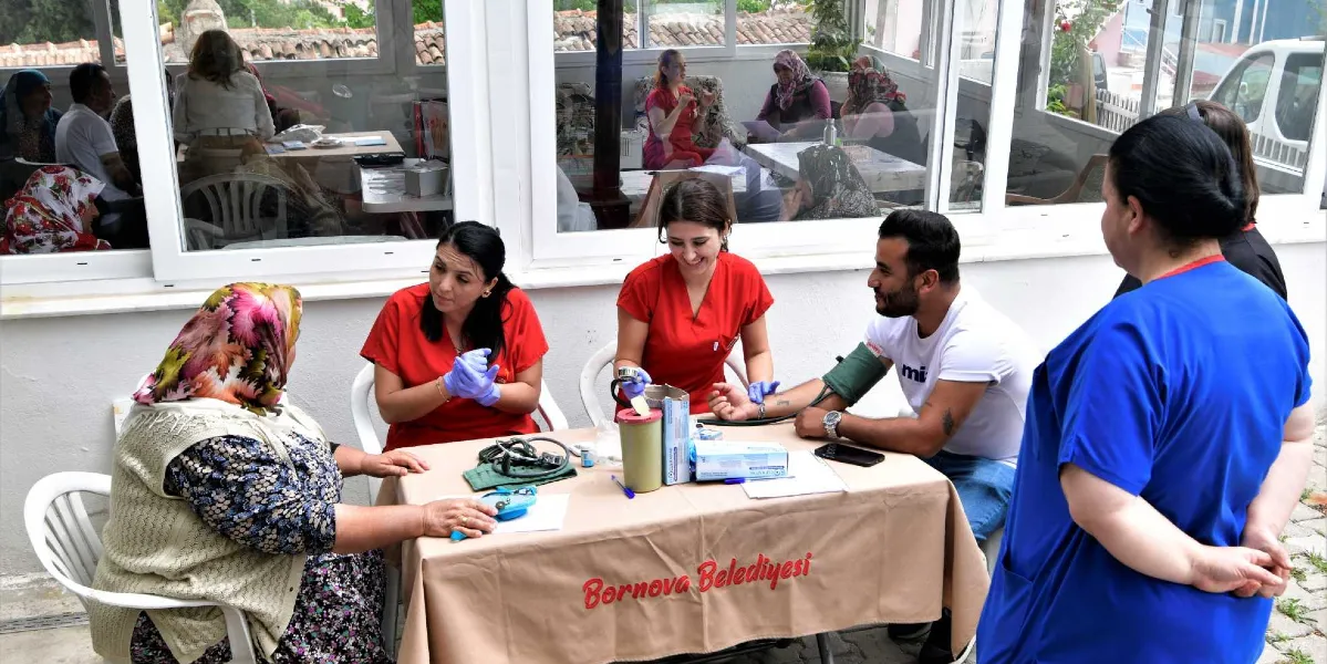 Bornova Belediyesi tarafından kurulan bir stantta, kırmızı üniformalı iki sağlık çalışanı köylü vatandaşlara sağlık hizmeti veriyor. Sağ taraftaki sağlık çalışanı genç bir adamın tansiyonunu ölçerken, sol taraftaki diğer çalışan ise karşısında oturan ve başında çiçekli bir yazma bulunan yaşlı bir kadınla ilgileniyor. Masanın üzerinde tansiyon aleti, eldiven kutusu ve tıbbi malzemeler bulunuyor. Arka planda camlı bir bölmenin içinde oturan diğer vatandaşlar da görülüyor.