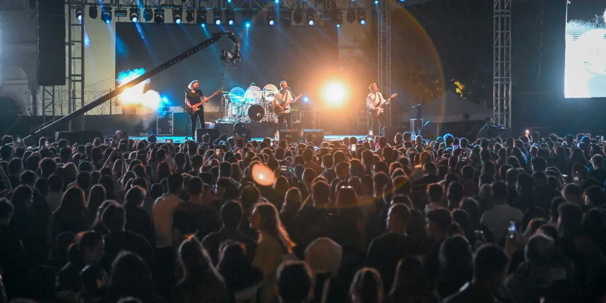 Gece vakti açık havada düzenlenen bir konserde, sahnedeki müzik grubu performans sergilerken önlerinde toplanan kalabalık bir izleyici kitlesi görülüyor. Sahne ışıklarıyla aydınlatılmış alanda bas gitarist, baterist, gitarist ve vokalist yer alıyor. Sahnenin arkasında büyük bir ekran ve profesyonel ışıklandırma sistemleri bulunurken, izleyiciler sahneye doğru dönmüş şekilde konseri izliyor.