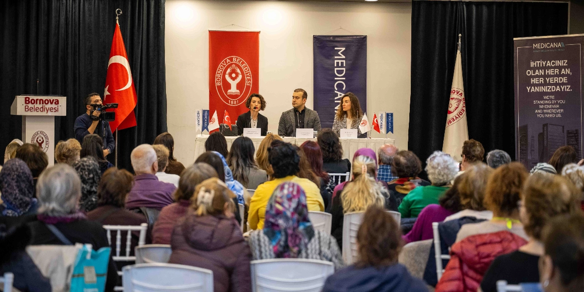 Bornova Belediyesi ve Medicana Sağlık Grubu iş birliğiyle düzenlenen farkındalık etkinliğinde, üç konuşmacı bir masada oturarak katılımcılara hitap ediyor. Salonun ön tarafında, arkalarında Türk bayrağı, Bornova Belediyesi ve Medicana logolu flamaların bulunduğu bir sahnede konuşmacılar yer alırken, izleyici koltuklarında oturan kalabalık bir grup onları dinliyor. Sahnenin sol tarafında bir kameraman çekim yapıyor, sağ tarafta ise üzerinde 