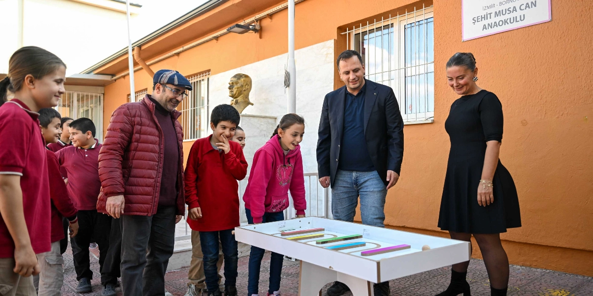 İzmir Bornova'daki Şehit Musa Can Anaokulu'nun bahçesinde, bir grup öğrenci ve yetişkin, üzerinde renkli parkurlar bulunan bir oyun masasının etrafında toplanmış eğleniyor. Çocuklar ve yetişkinler oyun masasına bakarak gülümserken, arka planda Atatürk büstü ve okul binasının turuncu renkli duvarı görülüyor.