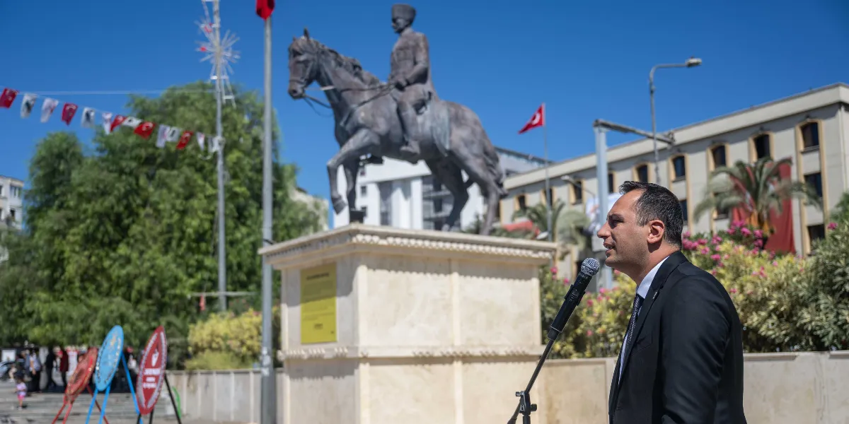 Bornova'da düzenlenen 9 Eylül anma töreninde, siyah takım elbiseli bir yetkili mikrofon başında konuşma yapıyor. Arka planda, üzerinde Atatürk'ün at üzerindeki heykelinin bulunduğu kaide ve çevresinde Türk bayraklarıyla süslenmiş bir meydan yer alıyor.